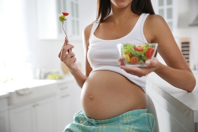 Photo of Young pregnant woman with bowl of vegetable salad at table in kitchen, closeup. Taking care of baby health