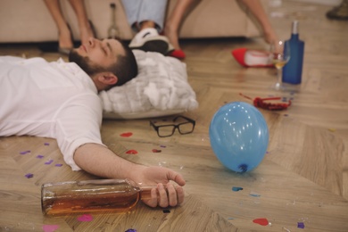 Photo of Man with bottle of wine sleeping on floor in messy room after New Year party