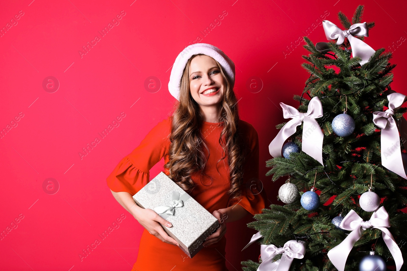 Photo of Beautiful young woman in Santa hat with gift box near Christmas tree on color background