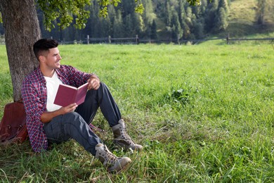 Photo of Handsome man reading book under tree on green meadow near forest