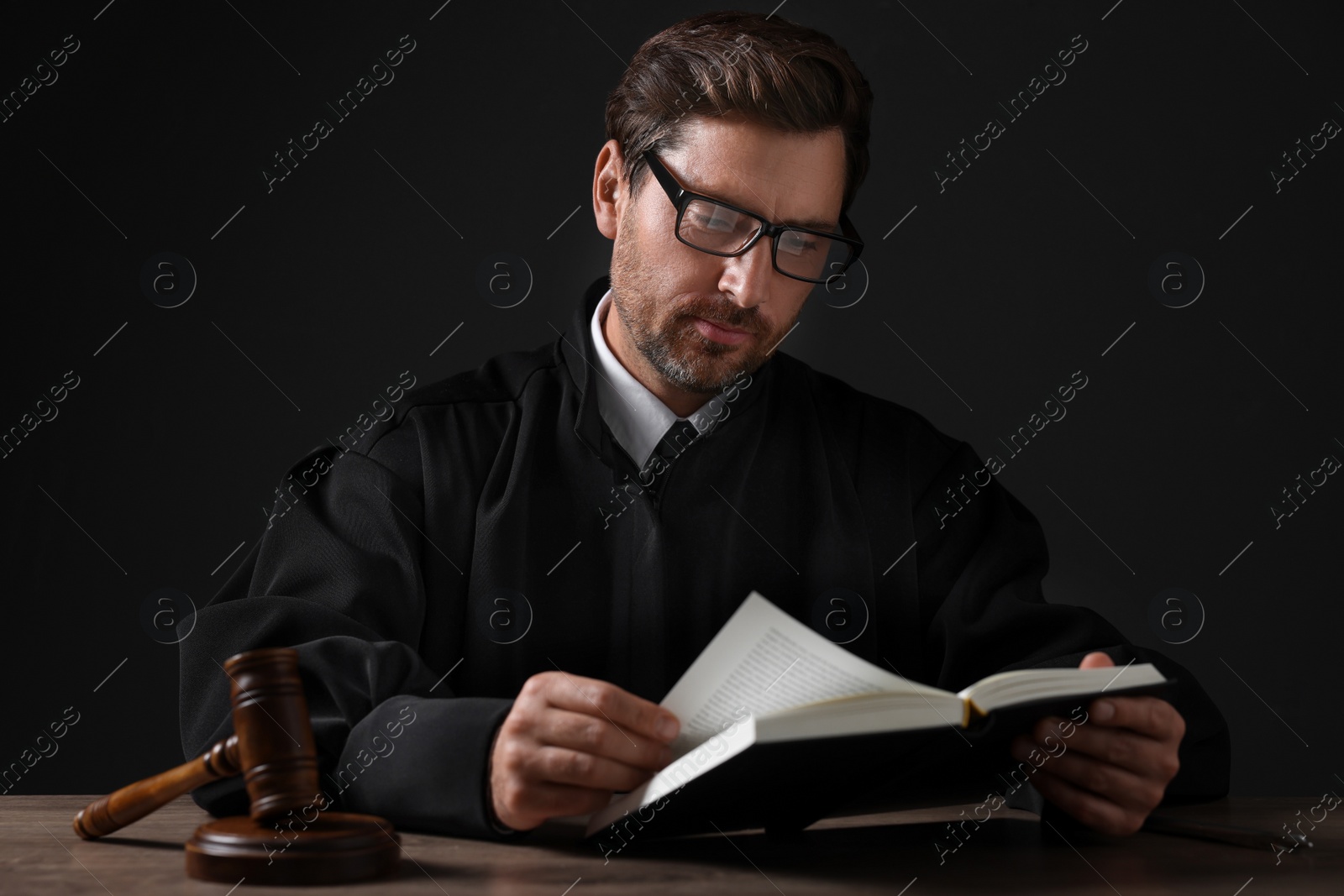 Photo of Judge with gavel reading book at wooden table against black background