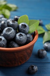 Photo of Tasty fresh blueberries with leaves in bowl on blue wooden table, closeup