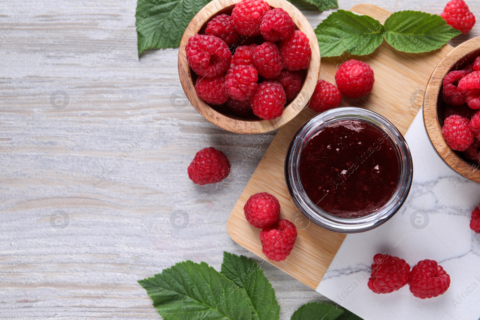 Photo of Delicious raspberry jam, fresh berries and green leaves on light wooden table, flat lay. Space for text