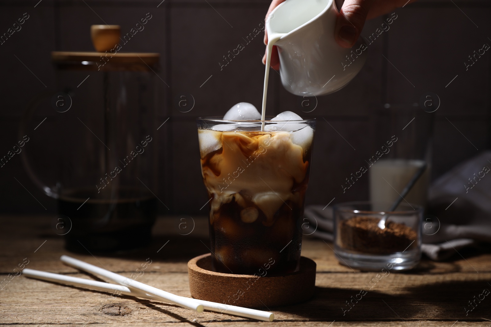 Photo of Woman pouring milk into glass with refreshing iced coffee at wooden table, closeup