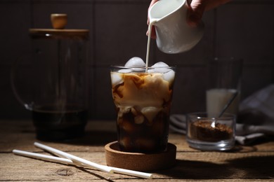 Photo of Woman pouring milk into glass with refreshing iced coffee at wooden table, closeup