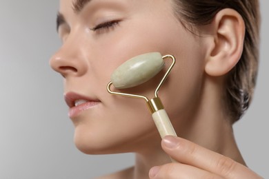 Young woman massaging her face with jade roller on grey background, closeup