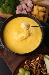 Photo of Dipping different products into fondue pot, fork with melted cheese on black wooden table, flat lay