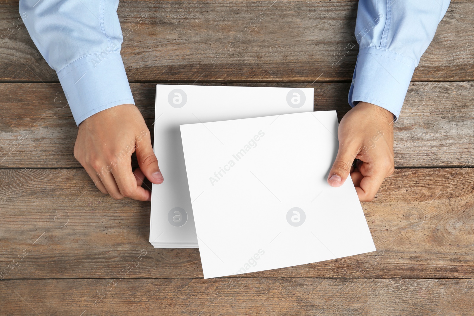 Photo of Man holding blank paper sheets for brochure at wooden table, top view. Mock up