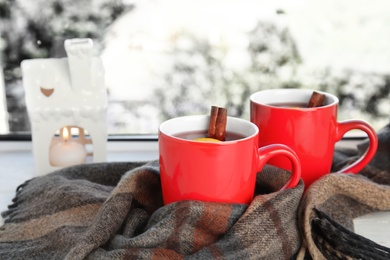 Photo of Cups of hot winter drink with scarf on window sill indoors