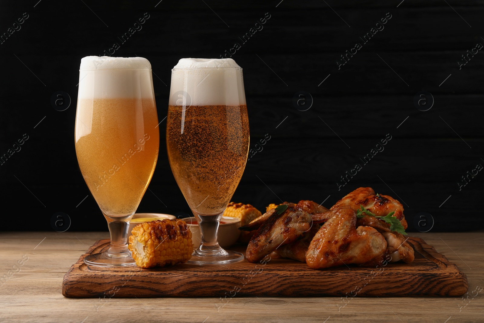 Photo of Delicious baked chicken wings, grilled corn and glasses with beer on wooden table against black background