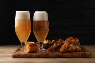 Delicious baked chicken wings, grilled corn and glasses with beer on wooden table against black background