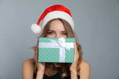 Happy young woman wearing Santa hat with Christmas gift on grey background