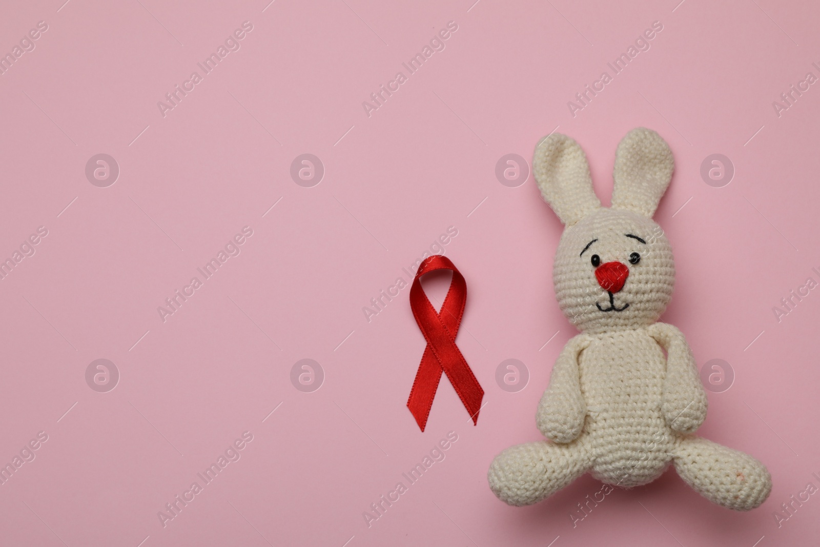 Photo of Cute knitted toy bunny and red ribbon on pink background, flat lay with space for text. AIDS disease awareness