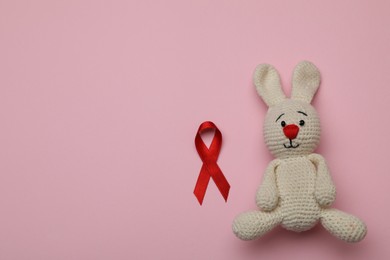 Photo of Cute knitted toy bunny and red ribbon on pink background, flat lay with space for text. AIDS disease awareness