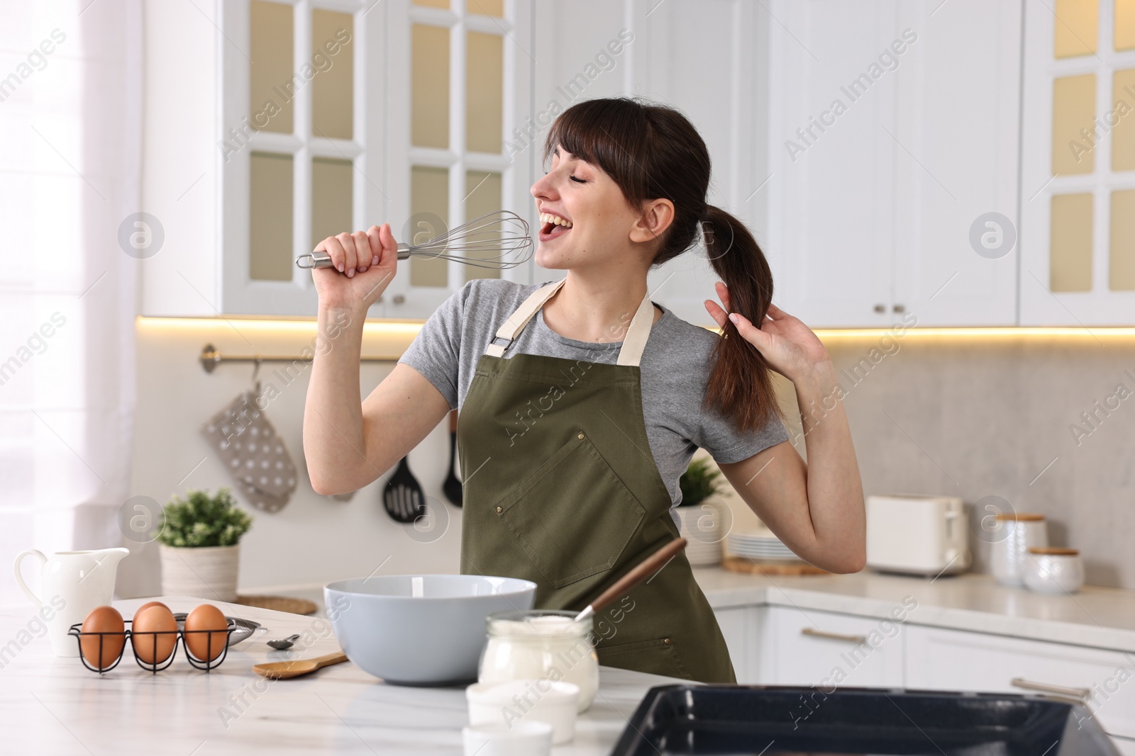 Photo of Happy young housewife with whisk having fun while cooking at white marble table in kitchen