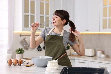 Happy young housewife with whisk having fun while cooking at white marble table in kitchen