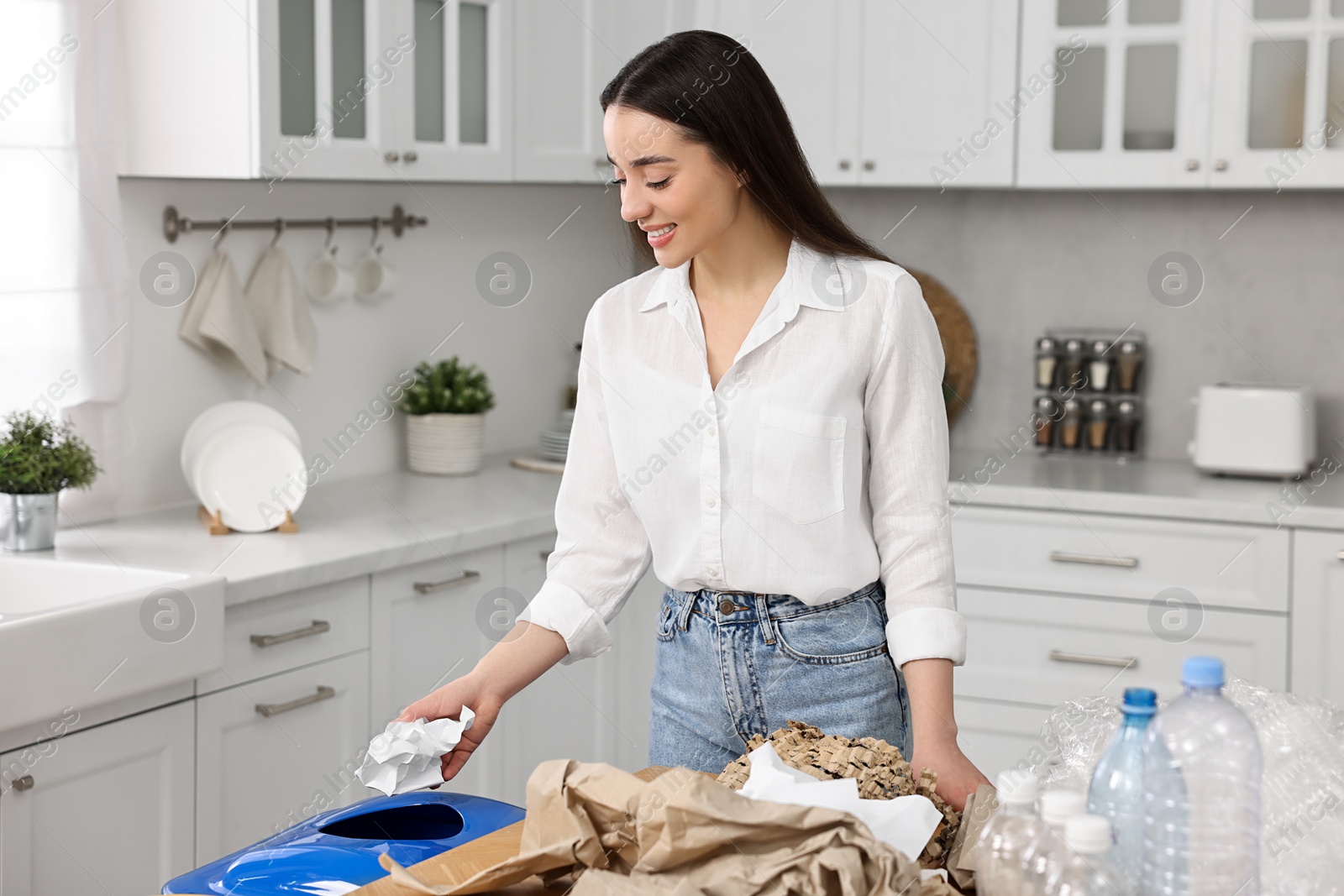 Photo of Garbage sorting. Smiling woman throwing crumpled paper into trash bin in kitchen