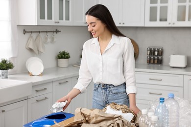 Photo of Garbage sorting. Smiling woman throwing crumpled paper into trash bin in kitchen