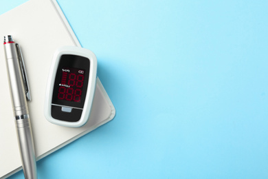 Photo of Modern fingertip pulse oximeter, pen and open notebook on light blue background, flat lay. Space for text