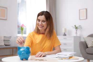 Photo of Woman putting coin into piggy bank at table in living room. Saving money