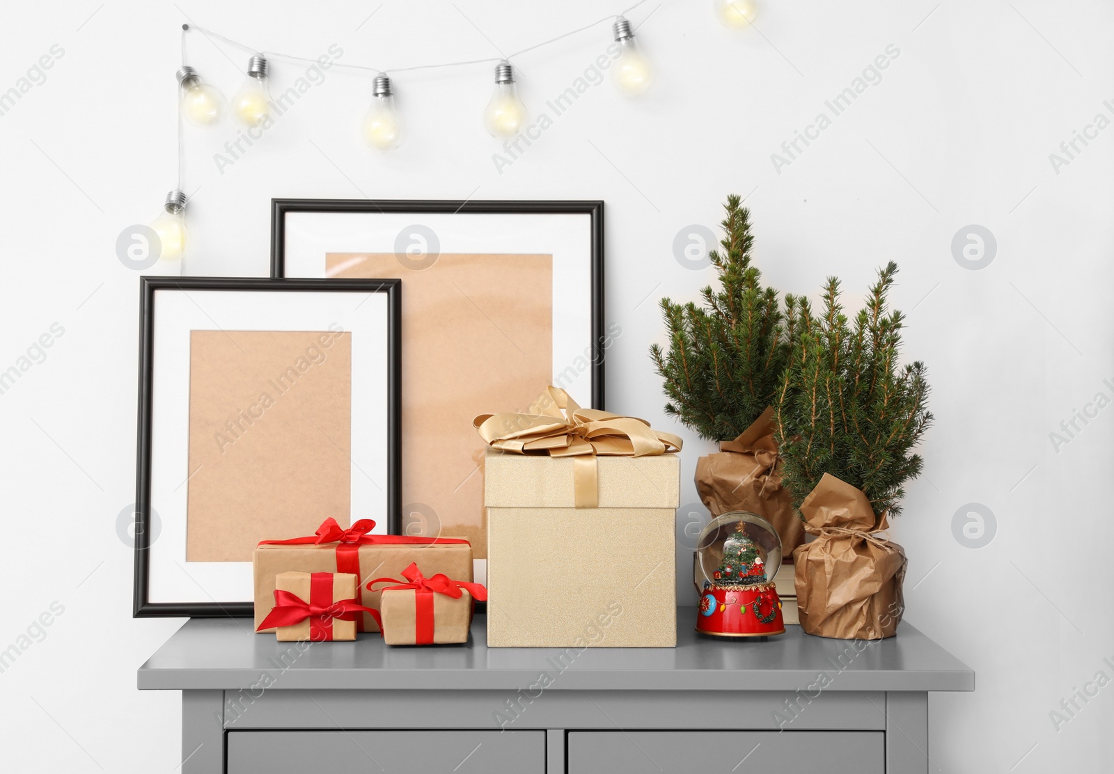 Photo of Empty frames, Christmas gifts, evergreen trees and accessories on grey chest of drawers indoors
