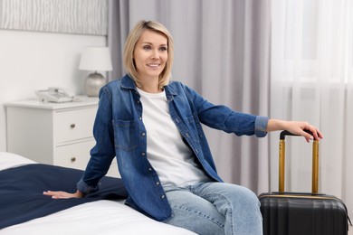 Photo of Smiling guest with suitcase relaxing on bed in stylish hotel room