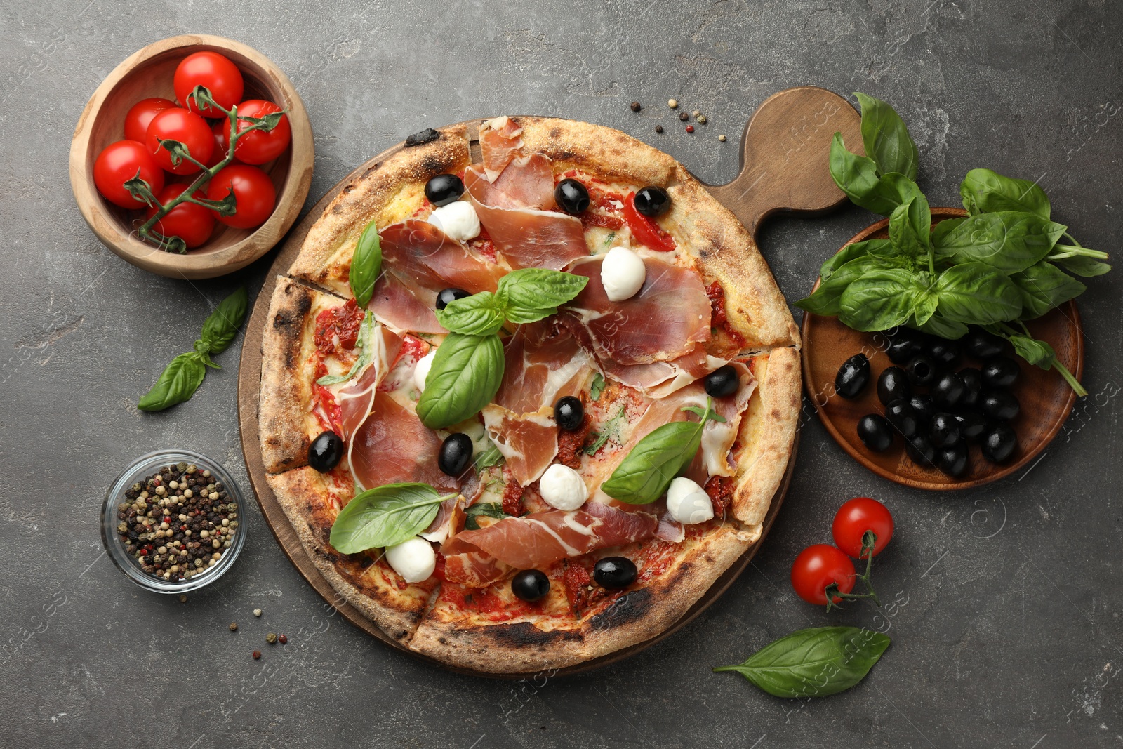 Photo of Tasty pizza with cured ham, olives, mozzarella cheese, tomatoes and arugula on gray table, flat lay