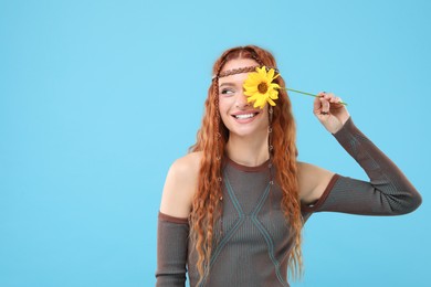 Photo of Beautiful young hippie woman covering eye with sunflower on light blue background, space for text