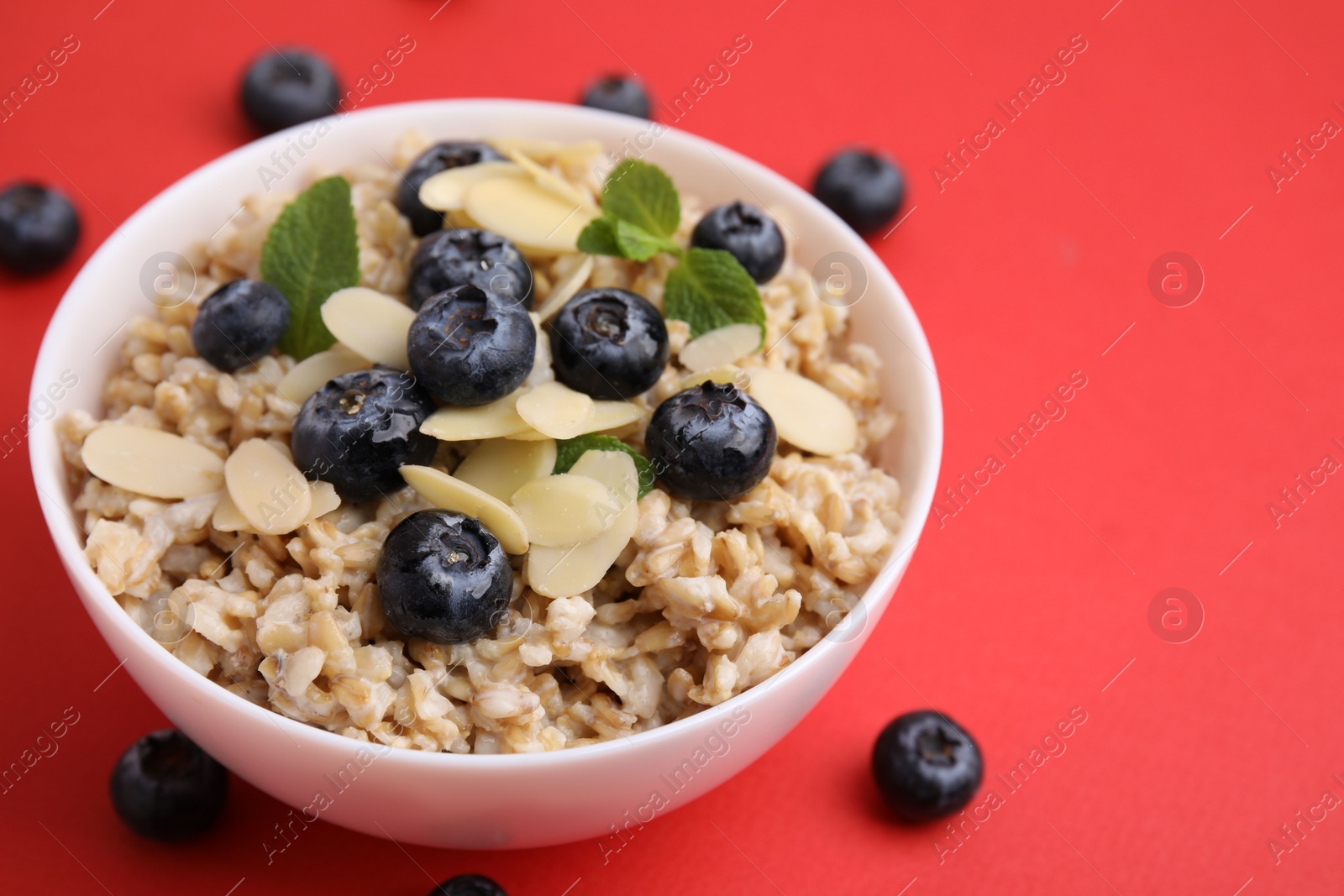 Photo of Tasty oatmeal with blueberries, mint and almond petals in bowl surrounded by fresh berries on red background, closeup. Space for text