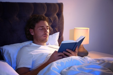 Photo of Handsome young man reading book in dark room at night. Bedtime