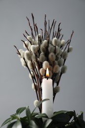 Photo of Burning candle, green leaves and willow branches against grey background