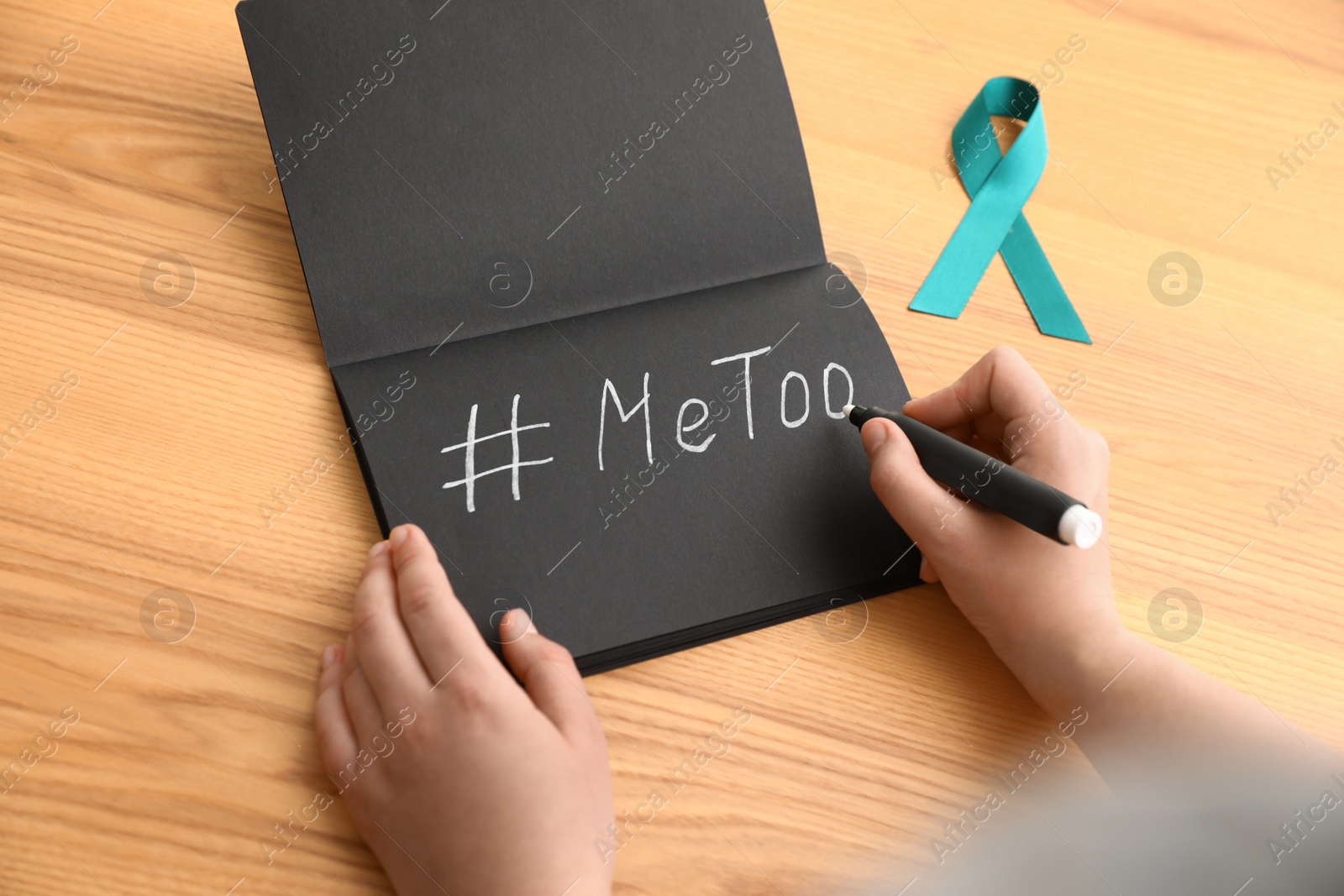 Photo of Woman writing hashtag MeToo in notebook near teal awareness ribbon at table, closeup. Stop sexual assault