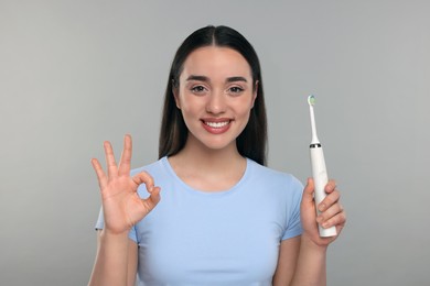 Photo of Happy young woman holding electric toothbrush and showing OK gesture on light grey background