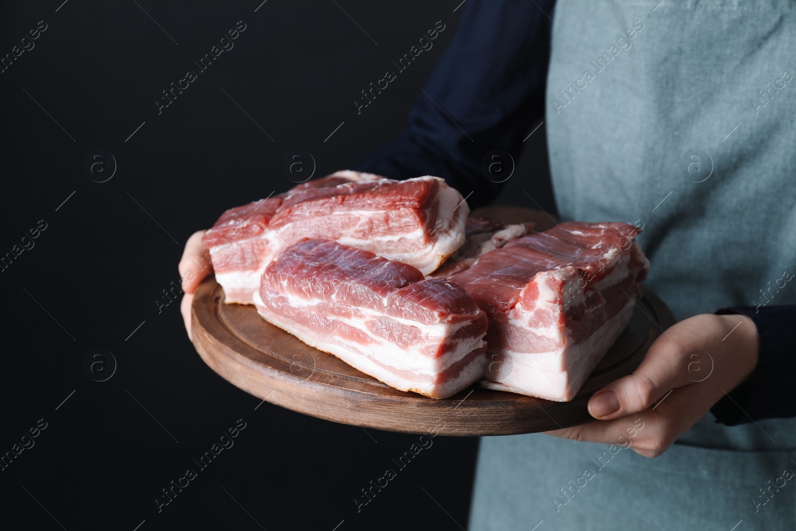 Photo of Woman holding wooden board with pieces of raw pork belly on black background, closeup