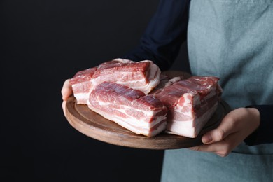 Woman holding wooden board with pieces of raw pork belly on black background, closeup