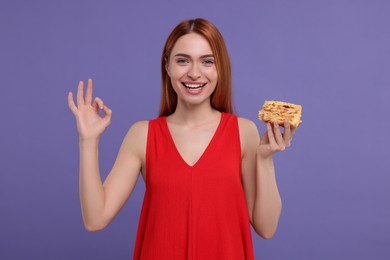 Photo of Young woman with piece of tasty cake showing ok gesture on purple background
