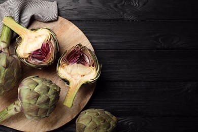 Photo of Cut and whole fresh raw artichokes on black wooden table, flat lay. Space for text