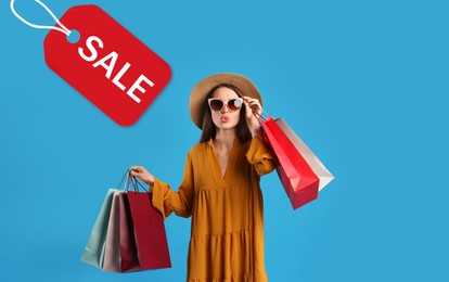 Image of Beautiful young woman holding paper shopping bags and tag with word SALE on light blue background