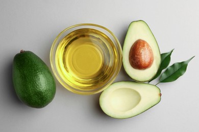 Photo of Cooking oil in bowl and fresh avocados on light grey background, flat lay