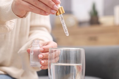 Photo of Woman dripping food supplement into glass of water indoors, closeup