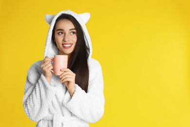 Young woman in bathrobe with cup of morning drink on yellow background. Space for text