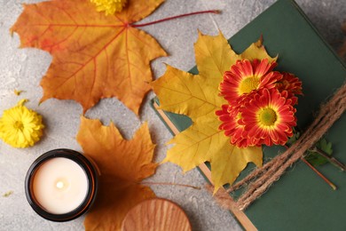 Book decorated with chrysanthemum flowers, autumn leaves and scented candle on light gray textured table, flat lay