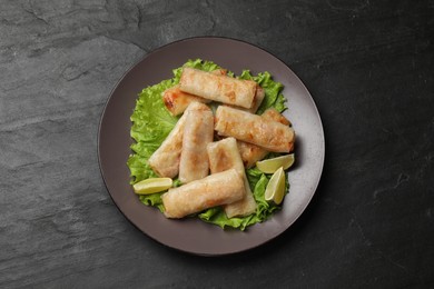 Plate with tasty fried spring rolls, lettuce and lime on dark textured table, top view