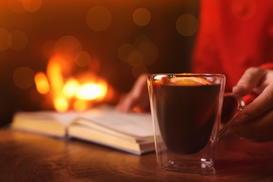 Woman with mulled wine reading book near fireplace indoors, closeup. Winter vacation