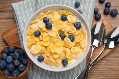 Photo of Bowl of tasty crispy corn flakes with milk and blueberries on wooden table, flat lay