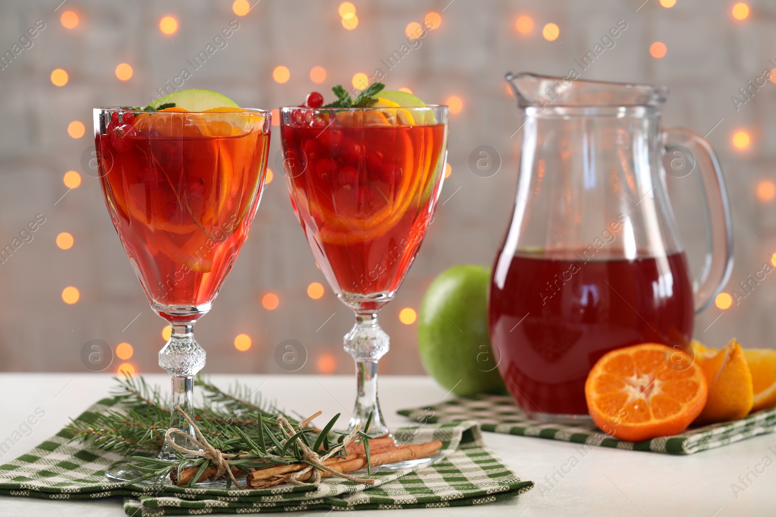 Photo of Christmas Sangria cocktail in glasses and jug, ingredients and fir tree branch on white table against blurred lights