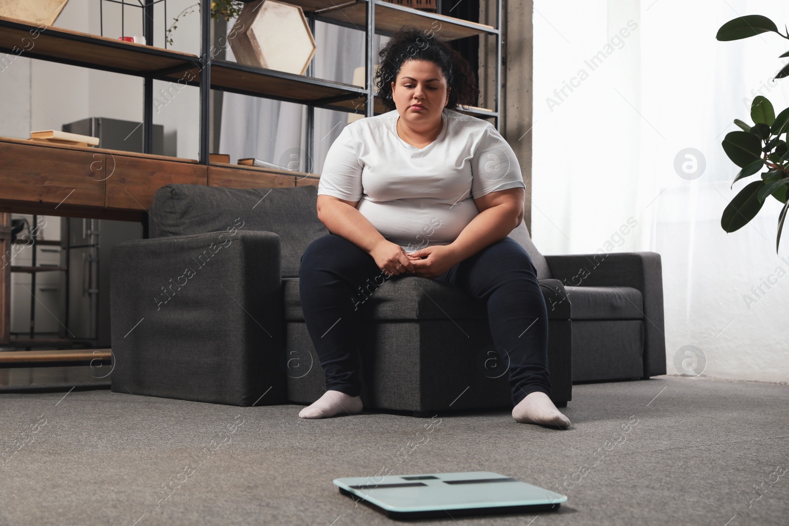 Photo of Depressed overweight woman looking at scales in living room