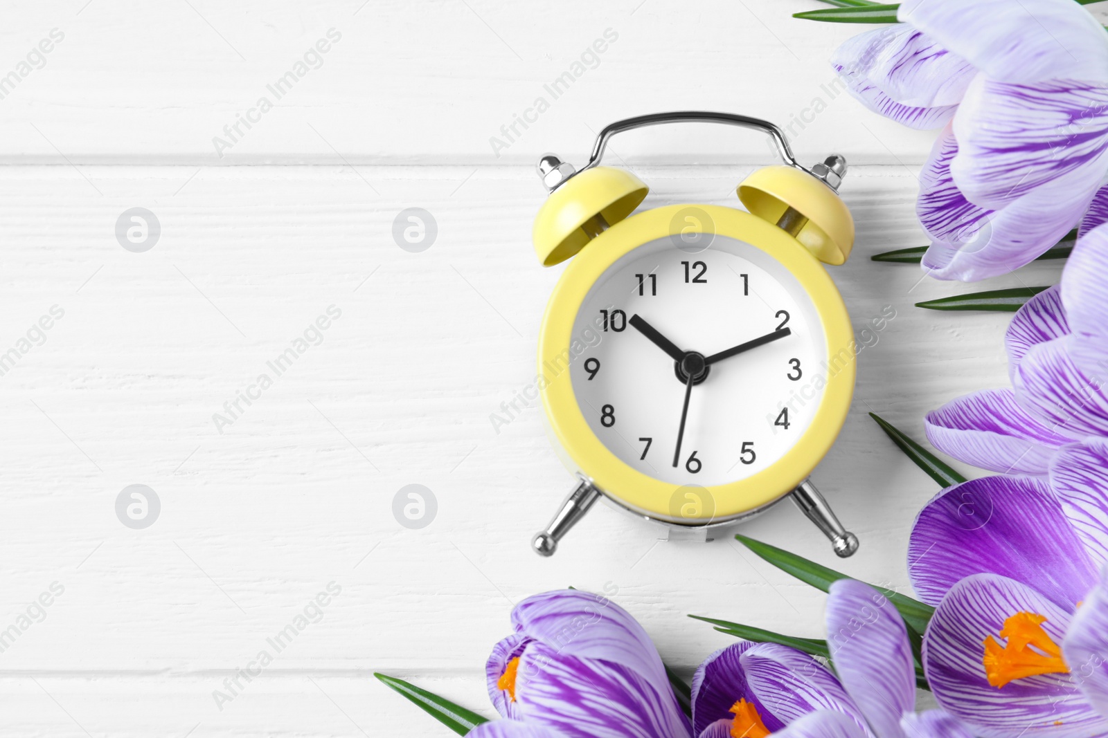 Photo of Yellow alarm clock, spring flowers and space for text on white wooden table, flat lay. Time change