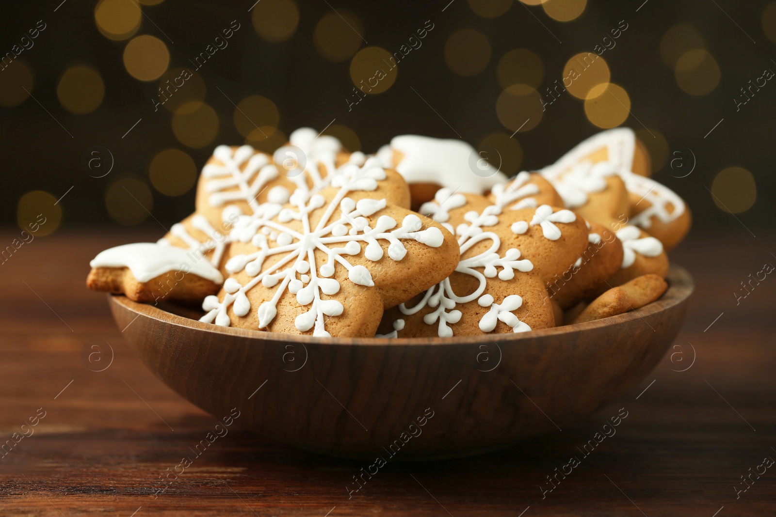 Photo of Tasty Christmas cookies with icing in bowl on wooden table against blurred lights, closeup