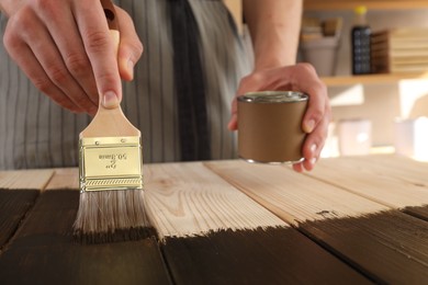 Man with brush and can applying wood stain onto wooden surface indoors, closeup